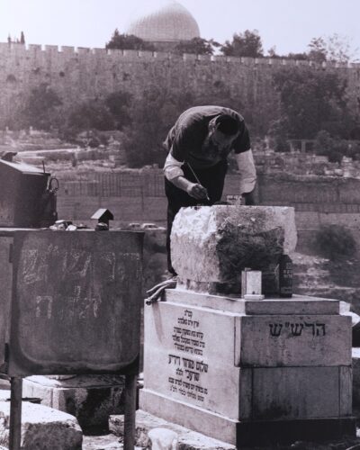 Analogue film Photography of a Jewish man painting Hebrew inscriptions on gravestones in Jerusalem, with the old city alls and dome in the background, by Jeroen Busschers