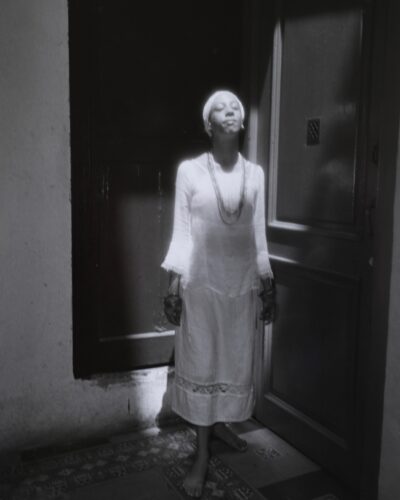 Analogue film photography portrait of a santera priestess in white dress standing in a sunbeam doorway, Santeria culture in Havana Cuba, by Jeroen Busschers