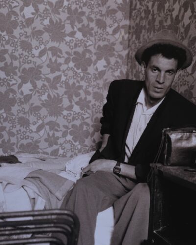 Analogue fil photography portrait of a caricature artist wearing a hat sitting on a bed in a French hotel room with retro floral wallpaper, black and white documentary style, by Jeroen Busschers